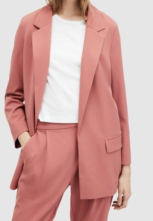 Femme portant un blazer rose poudré taillé sur mesure et un pantalon assorti, accompagnés d'un t-shirt blanc uni à col rond.