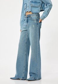 Ljust blå vida jeans i denim med en urblekt finish och subtila slitningar, kombinerade med en matchande kort denimjacka.