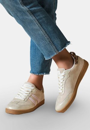 Pieds croisés portant des baskets beiges avec des lacets blancs, des rayures roses, un détail imprimé léopard et un jean bleu à ourlet effiloché.