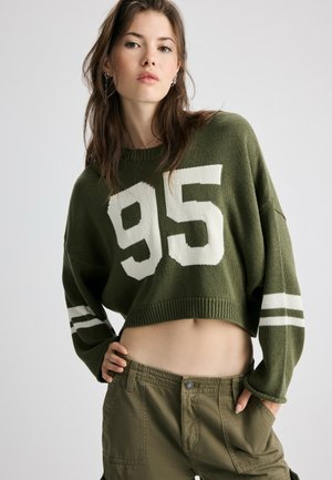 Jeune femme portant un pull court vert olive avec le numéro 95 et un pantalon cargo assorti, debout devant un fond uni.