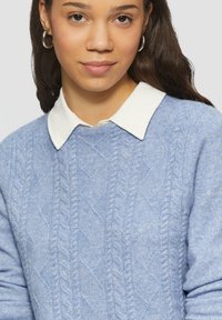Lichtblauwe gebreide trui met een gemêleerd patroon en kabelpatroon, laag over een witte blouse met kraag. Zilveren ringoorbellen zichtbaar.