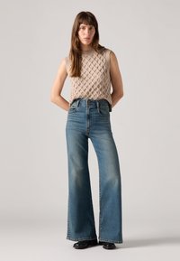 Top de malha sem mangas em bege com padrão texturizado, combinado com jeans flare de cintura alta azuis, apresentando um leve desbotamento e um clássico design de cinco bolsos.