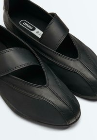 Chaussures noires à enfiler avec larges bandes élastiques, bouts pointus et dessus en cuir lisse, placées côte à côte sur un fond blanc.