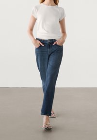 Witte shirt met korte mouwen en donkerblauwe jeans met een rechte pijl, gecombineerd met zwart-witte slippers, op een neutrale achtergrond.