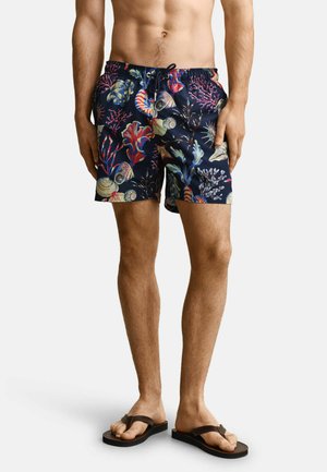 Torse et jambes d'un homme portant un short de bain bleu marine à motif coloré de coquillages et corail, associé à des tongs noires.