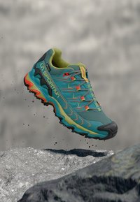 Trail running shoe with a turquoise upper, yellow accents, and a rugged black sole. Features textured mesh and a secure lace system.