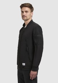 Schwarze Bomberjacke mit einem Frontreißverschluss, Stehkragen und seitlichen Taschen. Verfügt über ein strukturiertes Patch am Ärmeln und gerippte Bündchen.