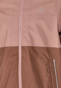 Vaaleanpunainen ja ruskea vetoketjullinen takki logolla. Ominaisuuksiin kuuluu sileä pinta, kaksi sivutaskua ja heijastavat yksityiskohdat.