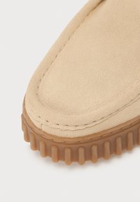 Beige suede shoe featuring a rounded toe design and a thick, ridged rubber sole. The stitching is visible along the edge.