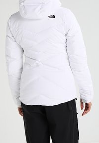 Weiße gepolsterte Jacke mit Kapuze, ausgestattet mit einem Zickzackdesign und schwarzem Logo auf der Rückseite. Tragen Sie sie mit schwarzen Hosen, glatter Textur.