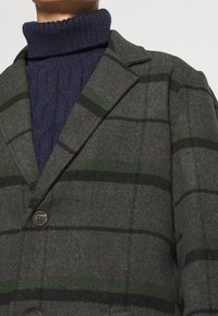 Grå tartan uldblazer med grønne og sorte striber, enkelt knaplukning og slagkrave, lagdelt over en mørkeblå kabelstrik turtleneck.
