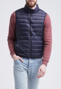 Námořnická puffer vesta s horizontálně prošívaným designem, vysokým límcem a předním zipem, nošená přes vínově červenou dlouhou rukávu. Na spodní části jsou vidět džíny.