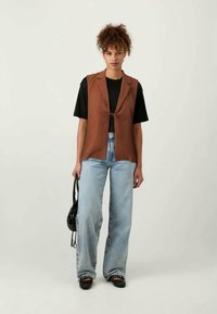 Gilet marrone senza maniche con dettaglio alla cravatta sopra una t-shirt nera, abbinato a jeans a gamba larga azzurri e scarpe nere. Borsa nera semplice.