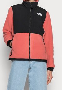 Osoba w dwukolorowej polarowej kurtce z czarną górą i koralowym dołem, zapięta, w połączeniu z jasnoniebieskimi dżinsami.