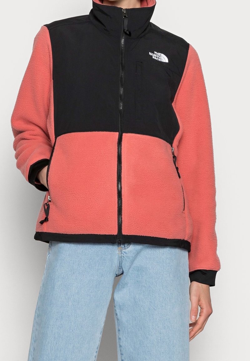 Osoba w dwukolorowej polarowej kurtce z czarną górą i koralowym dołem, zapięta, w połączeniu z jasnoniebieskimi dżinsami.