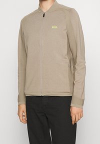 Světle hnědá zipová bunda s ribovanou texturou, vybavená vysokým límcem a malým zeleným logem "BOSS" na hrudi. Hladká tkanina a upravený střih.