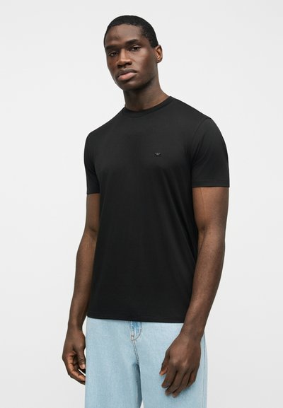 Άνδρας με σκούρο δέρμα που στέκεται, φοράει απλό μαύρο T-shirt και ανοιχτό μπλε τζιν, με φόντο μια απλή λευκή επιφάνεια.