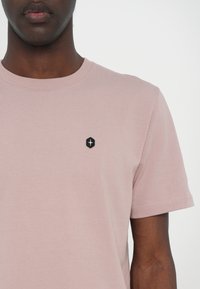 Helles rosa T-Shirt mit kurzen Ärmeln aus Baumwolle, versehen mit einem hexagonalen schwarzen Patch mit einem weißen Symbol auf der linken Brustseite.