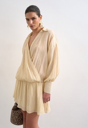 Γυναίκα με πίσω χτενισμένα μαλλιά φοράει πλισέ μπεζ φόρεμα με βαθύ V λαιμόκοψη και μακριά μανίκια, κρατώντας μια μικρή τσάντα με ρίγες ζέβρας.