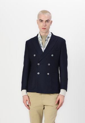 Blazer doppiopetto navy con bottoni argentati lucidi, due tasche frontali, rever a scollo e polsini in crema strutturata sotto.