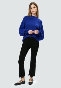 Blauer Strickpullover mit leicht schulterfreiem Design, kombiniert mit schwarzen Schlagjeans mit Spitzenbesatz und schwarzen Lackflats.