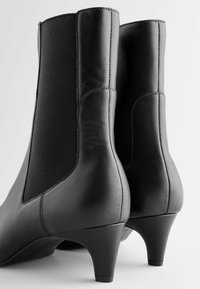 Bottines en cuir noir avec panneaux latéraux élastiques, bouts arrondis et talons effilés de 3 pouces. Texture lisse avec des éléments de design minimalistes.