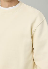 Sweatshirt de couleur crème avec un col ras du cou côtelé, une texture douce et des coutures d'épaule tombantes, montrant un tissu lisse sans motifs.