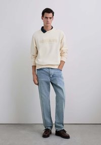 Joven vestido con un sudadero color crema con el texto "scalpers", jeans azules y zapatos marrón oscuro, de pie contra un fondo ligero y liso.