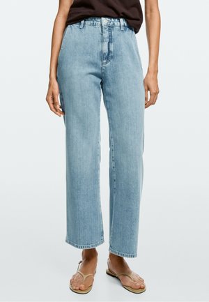 Jean bleu clair taille haute à jambes droites porté par une personne avec un haut noir et des sandales beiges, debout sur un fond clair uni.