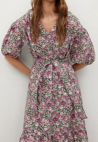 Robe imprimée florale avec manches bouffantes, décolleté en V et taille à nouer. Les motifs comprennent des fleurs roses et blanches sur un fond vert foncé.