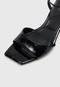 Sandal de tacón alto negro hecho de un material brillante y texturizado con punta cuadrada y una correa ancha. La plantilla es suave y contorneada.