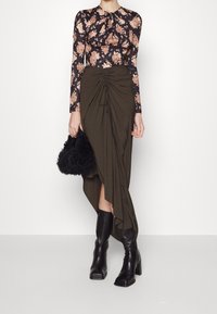 Blusa floreale a maniche lunghe con sfondo scuro, abbinata a una gonna verde oliva con dettagli drappeggiati e spacco laterale. Stivali neri al ginocchio e una borsa nera testurizzata.