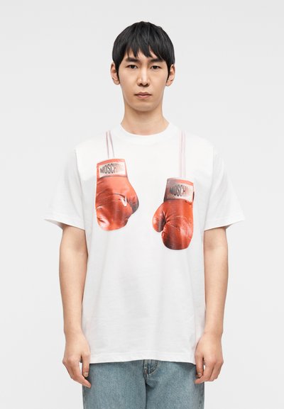 Λευκό βαμβακερό μπλουζάκι με ένα γραφικό που απεικονίζει κόκκινα γάντια πυγμαχίας. Τα γάντια έχουν μαύρο και λευκό λογότυπο και κρέμονται από μπεζ λουράκια.