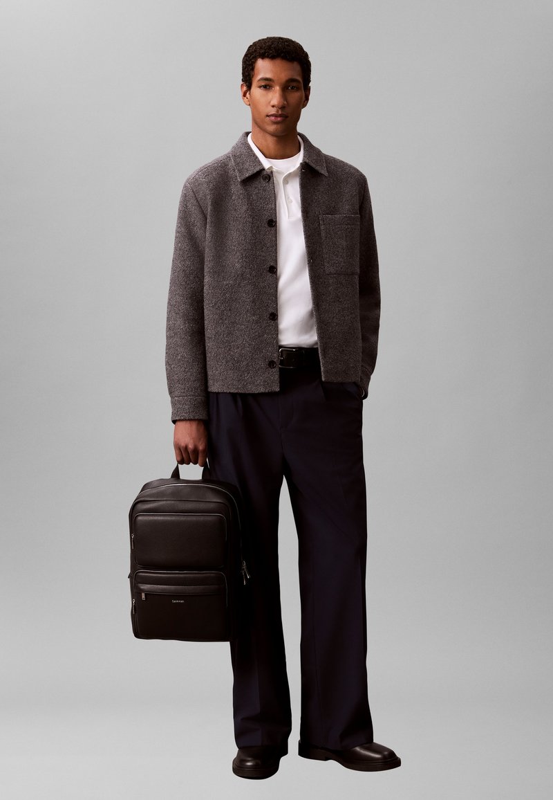 Jeune homme portant une veste grise boutonnée, une chemise blanche, un pantalon noir, tenant un sac à dos en cuir noir sur un fond gris uni.