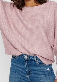 Ljust rosa, strukturerad tröja med vid halsringning, kombinerad med högmidjade, slitna blå jeans med en metallknapp och dragkedja.