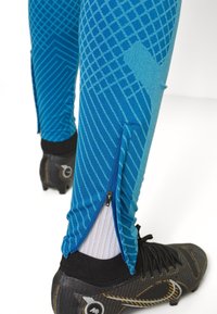 Blauwe sportlegging met geometrische patronen, ritssluitingen bij de enkels, gecombineerd met zwarte voetbalschoenen. Textuurstof met rek.