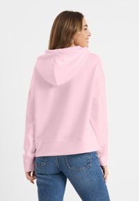 Lichtroze hoodie met een zachte textuur, voorzien van een capuchon met trekkoord en geribde boorden. Combinatie met blauwe denim jeans, gezien vanaf de achterkant.
