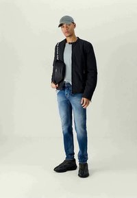 Melna bomber jaka, pelēks T-krekls, nolietoti zili džinsi, melni sporta apavi un pelēka cepure. Modelis tur melnu plecu somu ar baltu uzrakstu.