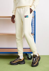 Lysegule sweatpants med sidelommer og Nike-logo patch. Sammen med sorte og brune sneakers med mesh-detaljer og synlig polstring.