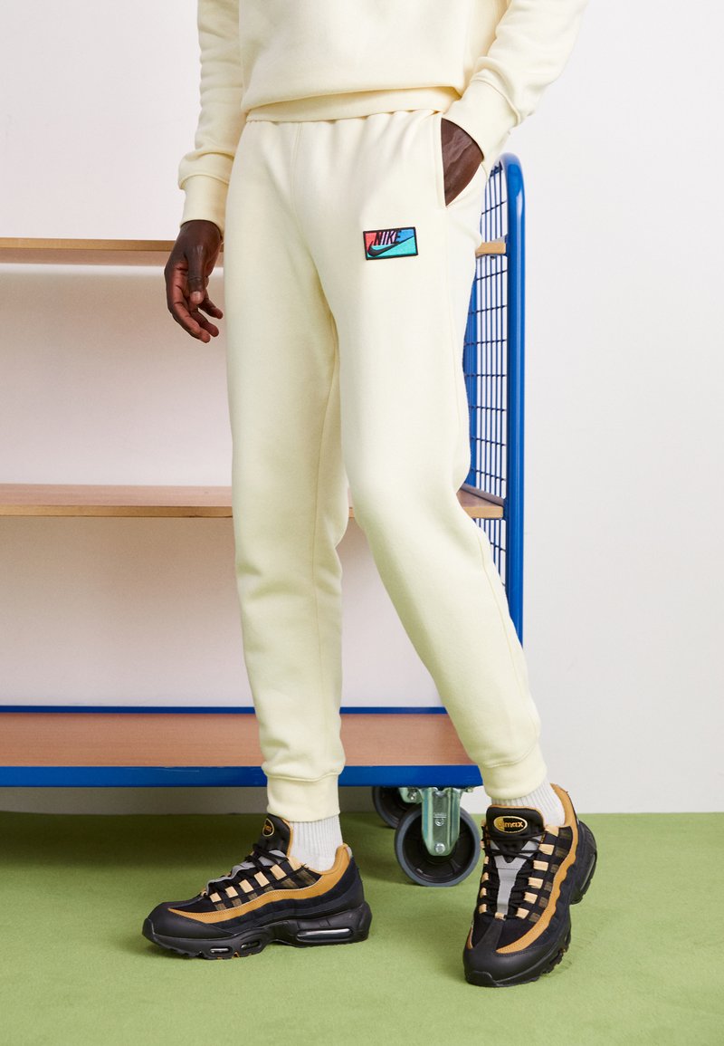Lysegule sweatpants med sidelommer og Nike-logo patch. Sammen med sorte og brune sneakers med mesh-detaljer og synlig polstring.