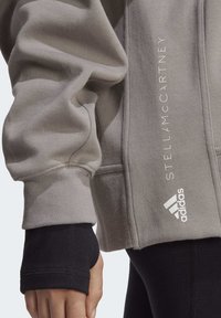 Sudadera gris con puños acanalados y bolsillos laterales. Presenta los logotipos blancos de "STELLAMCCARTNEY" y Adidas. Tejido suave y texturizado.