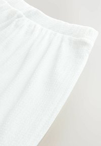 Bílá texturovaná tkanina s jemným vzorem, vybavená elastickým pasem. Material vypadá měkce a lehce.