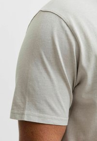Hellgraues Baumwoll-T-Shirt, das von der linken Schulter präsentiert wird, mit glatter Textur, kurzen Ärmeln und einem schlichten, klaren Design.