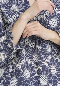 Blouse bleu marine avec broderies florales blanches, dotée de poignets à volants et de détails découpés complexes, présentant un motif délicat et texturé.