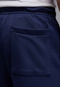 Mørkeblå sweats med struktureret stof, der har en åben baglomme. Elastisk talje synlig foroven. Enkel og funktionel design.