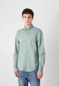 Lys grønn skjorte med knappelukking og brystlomme, teksturert stoff og krage. Brukt sammen med blå jeans, stående mot en ensfarget bakgrunn.