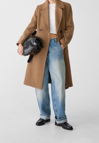 Personne portant un manteau marron, un crop top blanc, un jean large bleu clair, des mocassins noirs, tenant un sac à main noir brillant.