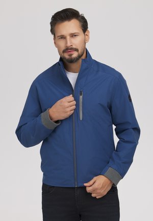 Mężczyzna w niebieskiej kurtce na zamek błyskawiczny z szarymi mankietami, trzymający zamek przy klatce piersiowej, stojący na tle jasnego, jednolitego tła.