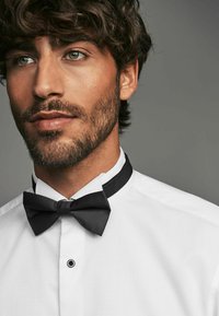 La camisa de cuello blanco presenta un tejido estructurado y texturizado con una pajarita negra. La pajarita tiene un sutil patrón de rayas y está atada de manera ordenada.