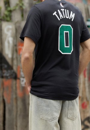 Person wearing a black Tatum basketball jersey with the number 0, standing against a graffiti-covered wall, hands in light blue jeans pockets.
